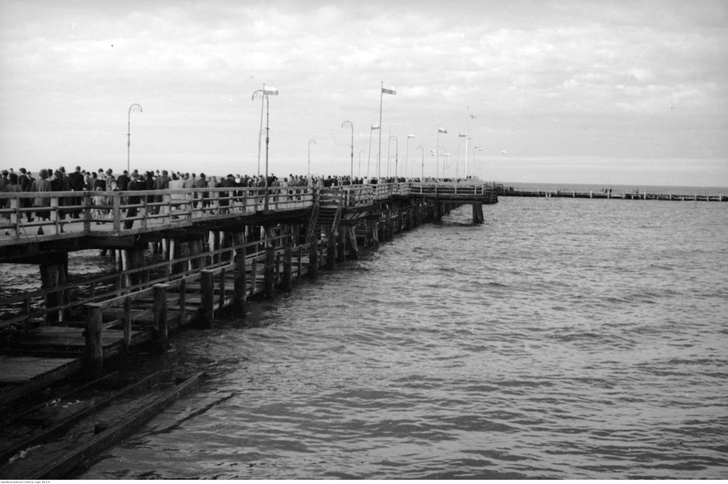 Molo - widok ogólny. Widoczne flagi państwowe i spacerujący ludzie. Fot. Narodowe Archiwum Cyfrowe. Molo - widok ogólny. Widoczne flagi państwowe i spacerujący ludzie. Fot. Narodowe Archiwum Cyfrowe.